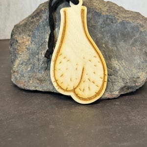 Puede incluir: Adorno de madera con forma de testículos, con detalles grabados y un cordón negro para colgar. El adorno es de madera clara con un acabado natural.