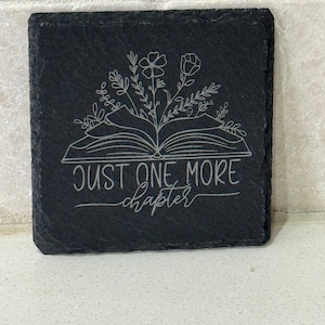 May include: Black slate coaster featuring a white design of an open book with flowers growing from it. The text "JUST ONE MORE chapter" is written beneath the book.