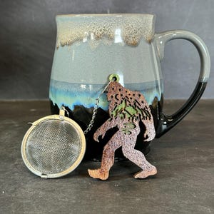 May include: A wooden Bigfoot tea infuser with a green mountain design. The infuser is hanging from a silver chain attached to a ceramic mug with a blue and gray glaze.