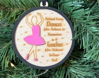 Puede incluir: Adorno navideño de madera con borde negro y una ilustración de bailarina rosa. El texto dice: "Detrás de cada bailarín que cree en sí mismo hay un profesor que creyó en ellos primero."