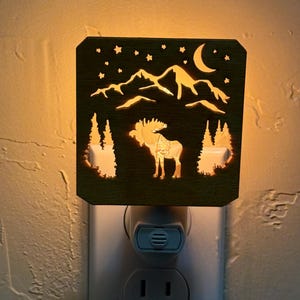 Op de afbeelding: Een houten nachtlampje met een uitgesneden ontwerp van een eland, bergen, sterren en een halve maan. Het nachtlampje is aangesloten op een wit stopcontact en geeft een warme, zachte gloed. Het ontwerp is een silhouet van een wildernislandschap.