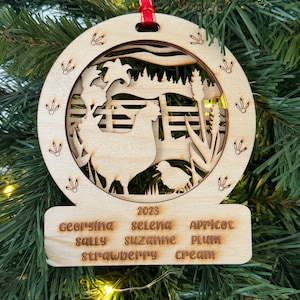 May include: A wooden Christmas ornament with a cutout design of a hen and chicks in a farmyard setting. The ornament is decorated with the year 2023 and the names Georgina, Sally, Selena, Suzanne, Apricot, Plum, Strawberry, and Cream.