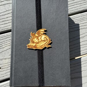 May include: A black book with a black elastic bookmark. The bookmark has a wooden charm attached to it. The charm is a dragon curled up on a book.