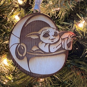 May include: A wooden Christmas ornament featuring a cartoon character from the Star Wars franchise, The Mandalorian. The character is depicted in a brown and white spaceship, with a light brown background.