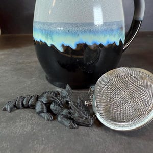 May include: A black dragon tea infuser with a silver mesh tea ball. The dragon is lying on a dark grey surface. The tea ball is attached to a chain.