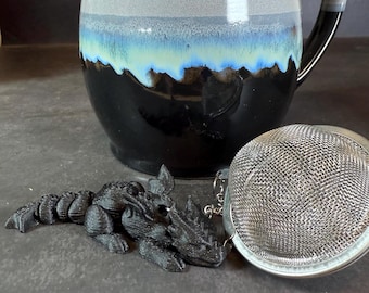 May include: A black dragon tea infuser with a silver mesh tea ball. The dragon is lying on a dark grey surface. The tea ball is attached to a chain.