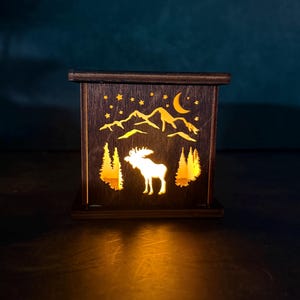 May include: A wooden lantern with a cutout design featuring a moose, trees, mountains, stars, and a crescent moon. The lantern is illuminated from within, casting a warm glow. The dark brown wood contrasts with the bright yellow light.