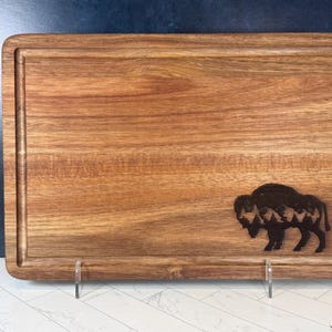 May include: Rectangular wooden cutting board with a carved bison silhouette. The board has a juice groove around the edge and is made of light brown wood. The bison design features a mountain scene within its form.
