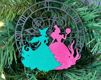 Puede incluir: Un adorno navideño de madera negro con forma de reloj con una silueta de dos mujeres en su interior. Una mujer lleva un vestido verde y la otra lleva un vestido rosa.