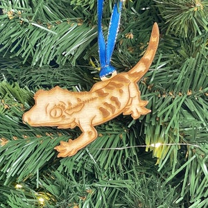May include: A wooden Christmas ornament shaped like a gecko lizard with a blue ribbon attached. The ornament is painted with brown and white stripes.