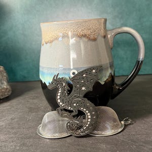 May include: A ceramic mug with a gray, blue, and black glaze, paired with a dragon-shaped tea infuser. The infuser is black with star cutouts. The mug has a large handle and is approximately 4 inches tall.