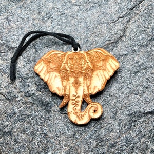 Puede incluir: Adorno de elefante de madera con detalles tallados. El adorno marrón claro presenta la cabeza y la trompa de un elefante, con un cordón negro para colgar. La cabeza del elefante está adornada con patrones florales y geométricos.