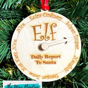 May include: Wooden Christmas ornament shaped like a compass with a spinning arrow. The ornament is decorated with the words "Extra-Ordinary", "Super-Duper", "Naughty", "Zone", "Not Very Nice", "Nice", and "Elf". The ornament also includes the text "Daily Report To Santa".