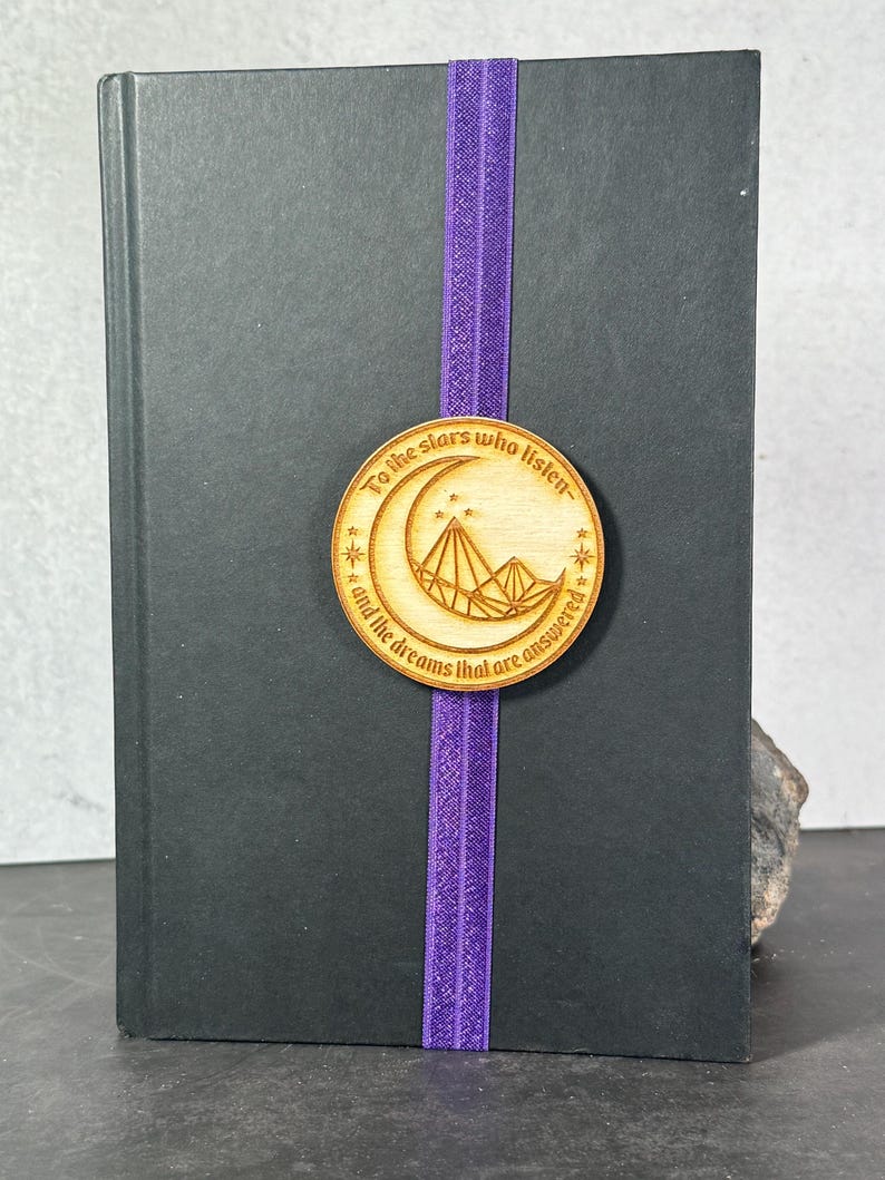 May include: A wooden bookmark with a crescent moon and mountain design. The bookmark has the text "To the stars who listen and the dreams that are answered".