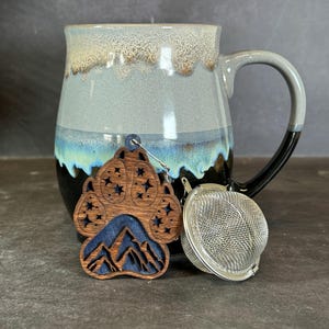 May include: A ceramic mug with a blue and brown glaze, a wooden paw print tea infuser with a mountain design, and a stainless steel tea ball. The paw print has star cutouts and a blue mountain scene.