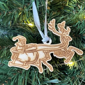 以下が含まれることがあります： 骨格のトナカイがサンタクロースの小さな人形を乗せたそりを引いている木製クリスマスオーナメント。オーナメントは茶色系の色で塗られており、素朴でヴィンテージな外観です。