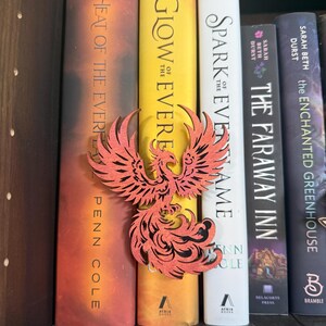 May include: A red phoenix-shaped wooden bookmark rests between colorful books on a shelf. The books have titles like "Heat of the Ever," "Glow of the Ever," and "Spark of the Evername." The bookmark adds a decorative touch to the book collection.