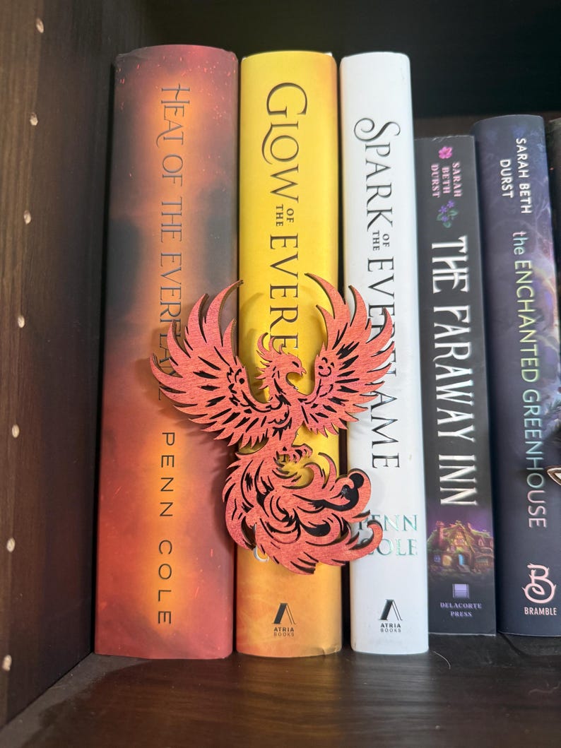 May include: A red phoenix-shaped wooden bookmark rests between colorful books on a shelf. The books have titles like "Heat of the Ever," "Glow of the Ever," and "Spark of the Evername." The bookmark adds a decorative touch to the book collection.