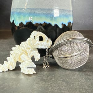 May include: A white ceramic dinosaur tea infuser with a silver mesh ball tea infuser attached to it by a chain.