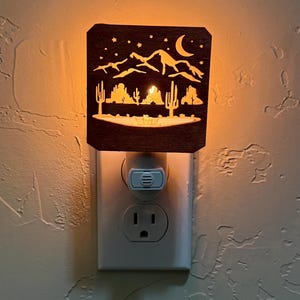 Peut inclure: Veilleuse en bois avec un motif de paysage désertique. La veilleuse présente un motif découpé de montagnes, de cactus, d'étoiles et d'un croissant de lune. La lumière brille de l'intérieur, créant une ambiance chaleureuse. La veilleuse est branchée sur une prise électrique standard.
