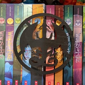 Op de afbeelding: Een zwart draakvormig bladwijzer staat voor een rij kleurrijke boeken. De boeken maken deel uit van de "Wings of Fire"-serie, met titels zichtbaar op de ruggen. De bladwijzer voegt een decoratief tintje toe aan de boekencollectie.