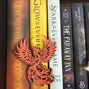 May include: A wooden phoenix book mark in front of a row of books. The phoenix is reddish-orange with intricate cut-out details. The book spines are various colors, including yellow, white, and black. Titles include "Heat of the Ever", "Glow of the Ever", and "Spark Everflame".