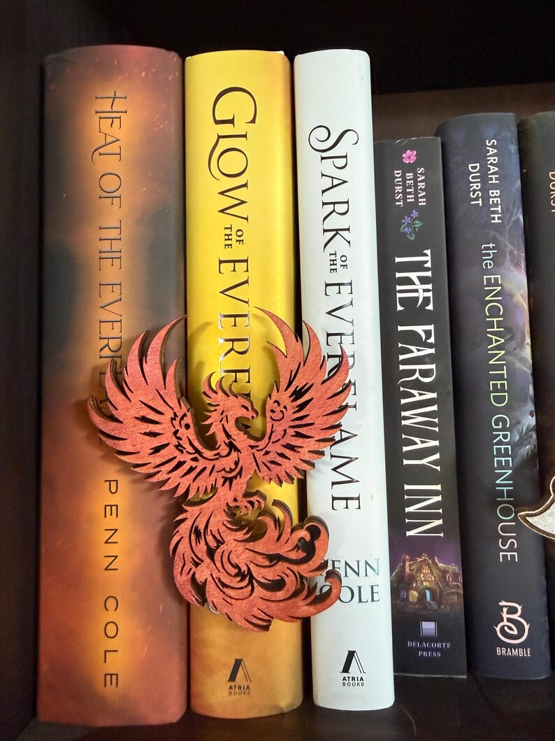 May include: A wooden phoenix book mark in front of a row of books. The phoenix is reddish-orange with intricate cut-out details. The book spines are various colors, including yellow, white, and black. Titles include "Heat of the Ever", "Glow of the Ever", and "Spark Everflame".