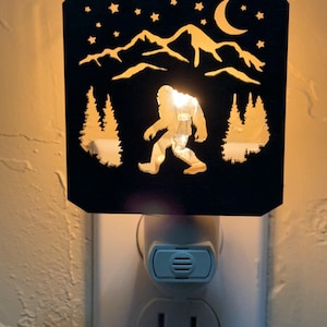 May include: Black night light featuring a cutout design of Bigfoot walking in front of mountains, trees, stars, and a crescent moon. The night light plugs into a standard wall outlet and emits a warm glow.