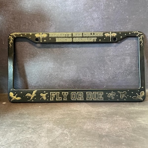 Op de afbeelding: Zwarte metalen kentekenhouder met gouden letters. De houder heeft de tekst "Baseaith War College Riders Quadrant" en "Fly or Die".