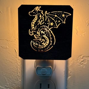 Puede incluir: Una luz de noche negra con un diseño de dragón dorado. El dragón tiene alas detalladas y un patrón de estrellas. La luz de noche se enchufa a una toma de corriente estándar, proporcionando un brillo suave y ambiental.