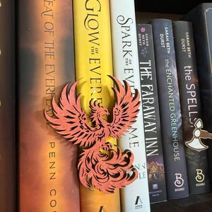 May include: A wooden phoenix bookmark in shades of orange and red rests among a row of books. The books have titles like "Glow of the Ever", "Spark of the Ever", and "The Faraway Inn". The bookmark is placed between the pages of a book.