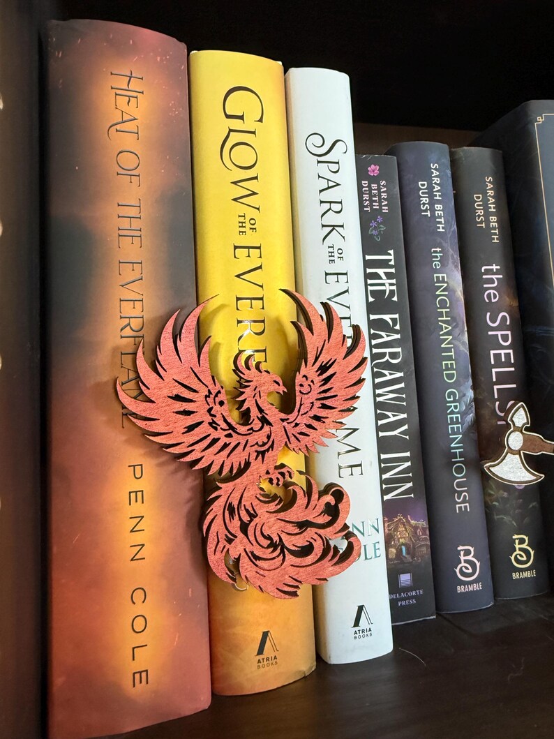 May include: A wooden phoenix bookmark in shades of orange and red rests among a row of books. The books have titles like "Glow of the Ever", "Spark of the Ever", and "The Faraway Inn". The bookmark is placed between the pages of a book.