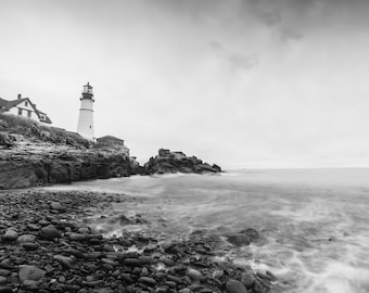 May include: A black and white photograph of a lighthouse on a rocky coastline. The lighthouse is tall and white with a black roof. The water is calm and the sky is cloudy.