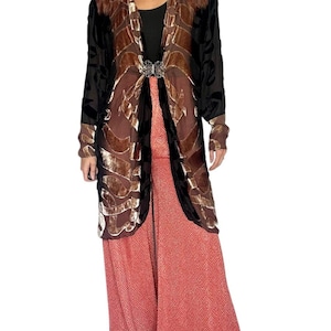 May include: A long-sleeved, open-front duster cardigan featuring a brown and black velvet burnout pattern, secured with a silver-toned metal clasp. Underneath is a black top paired with wide-leg, coral-colored trousers with a geometric pattern.