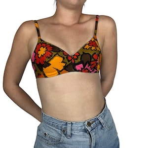 May include: A floral print bikini top with adjustable straps. The top features a colorful pattern of orange, red, and pink flowers on a green background. The bra is a triangle style with a V-neckline. The model is wearing blue denim jeans.