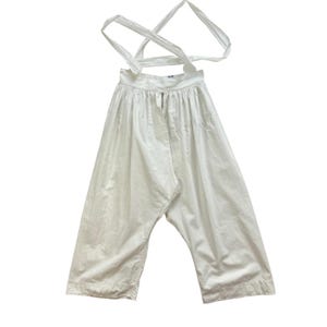 May include: A pair of white cotton trousers featuring a high-waisted design with gathered fabric at the waistband and long, thin shoulder straps that cross over the back. The pants have a wide-leg silhouette and a simple hemline at the bottom.