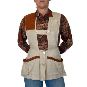 May include: A beige vest with a quilted brown shoulder patch and brown trim on the pockets, worn over a long-sleeved shirt featuring a brown, cream, and black geometric pattern. The vest has a button-front closure and multiple utility pockets.