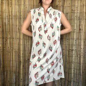 May include: A sleeveless, off-white knee-length dress featuring a repeating geometric pattern in red, teal, and black. The garment has a stand-up collar and a V-shaped neckline opening, with a straight, relaxed silhouette and a smooth, lightweight fabric.