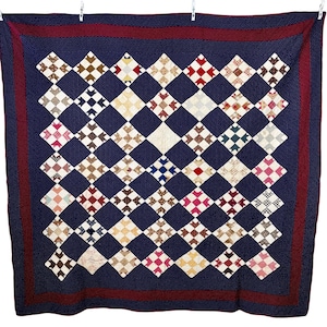 May include: A handmade quilt with a navy blue background featuring a repeating diamond pattern. The diamonds are filled with various patterned fabrics in shades of brown, red, white, and blue. A dark red border frames the quilt.