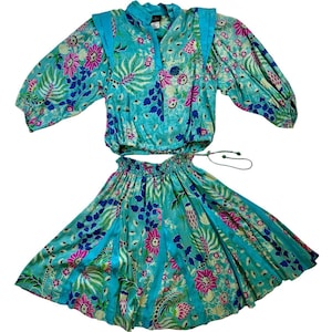 May include: A two-piece set consisting of a long-sleeved top and a skirt in a vibrant turquoise fabric featuring a multi-coloured floral and botanical print. The top has a V-neckline and shoulder accents, while the skirt features an elasticated waistband.