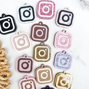 May include: A collection of square charms shaped like the Instagram logo, featuring various finishes including glitter, matte, metallic, and pearlescent textures in colors such as black, white, pink, gold, brown, purple, and lavender.