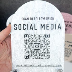 Może przedstawiać: Biały akrylowy znak z marmurowym wzorem, z kodem QR i tekstem "SCAN TO FOLLOW US ON SOCIAL MEDIA" oraz "www.millenniumhardwood.com". Znak jest trzymany przez dłoń.