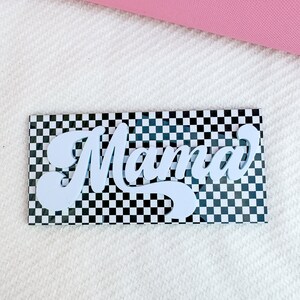 Pu&ograve; includere: Lettere bianche "Mama" in un font retr&ograve; su uno sfondo a scacchi bianco e nero.