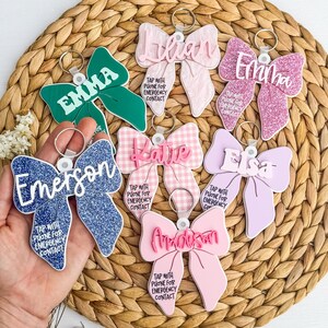 May include: A selection of bow-shaped keyrings in shades of green, pink, blue, and lilac. Each keyring displays a name and the text "TAP WITH PHONE FOR EMERGENCY CONTACT". The keyrings are set against a woven background.