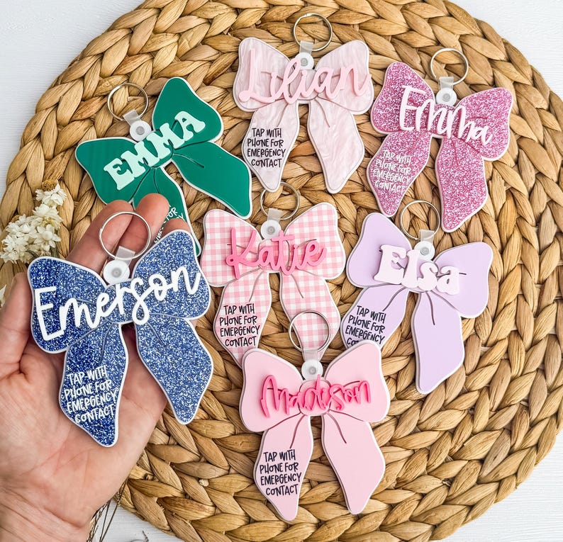 May include: A selection of bow-shaped keyrings in shades of green, pink, blue, and lilac. Each keyring displays a name and the text "TAP WITH PHONE FOR EMERGENCY CONTACT". The keyrings are set against a woven background.