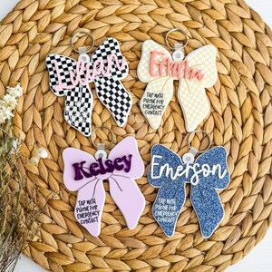 May include: Four bow-shaped keyrings with names and the text "TAP WITH PHONE FOR EMERGENCY CONTACT." The keyrings are in various colours and patterns, including checkered, glitter, and solid colours. Each keyring has a silver ring.
