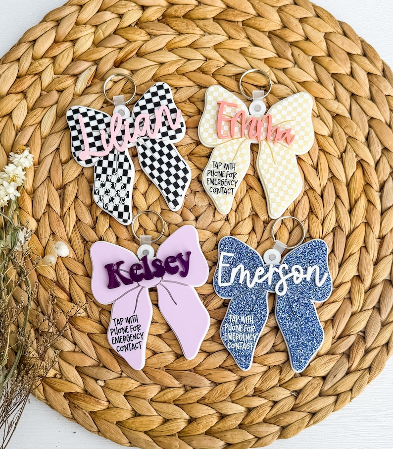 May include: Four bow-shaped keyrings with names and the text "TAP WITH PHONE FOR EMERGENCY CONTACT." The keyrings are in various colours and patterns, including checkered, glitter, and solid colours. Each keyring has a silver ring.