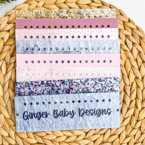 May include: A stack of colorful acrylic bands in various shades of pink, blue, and glitter. Each band has evenly spaced holes. The bottom band is light blue with the text "Ginger Baby Designs" in dark blue script.