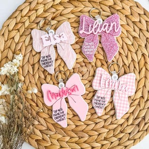 May include: Four pink bow-shaped keyrings with names and the text "TAP WITH PHONE FOR EMERGENCY CONTACT." One bow is glittery, one is gingham, and two are solid pink. Each keyring has a silver ring.