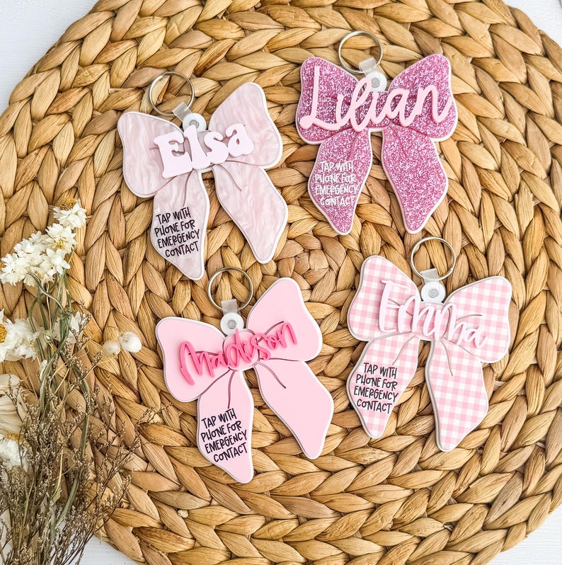 May include: Four pink bow-shaped keyrings with names and the text "TAP WITH PHONE FOR EMERGENCY CONTACT." One bow is glittery, one is gingham, and two are solid pink. Each keyring has a silver ring.